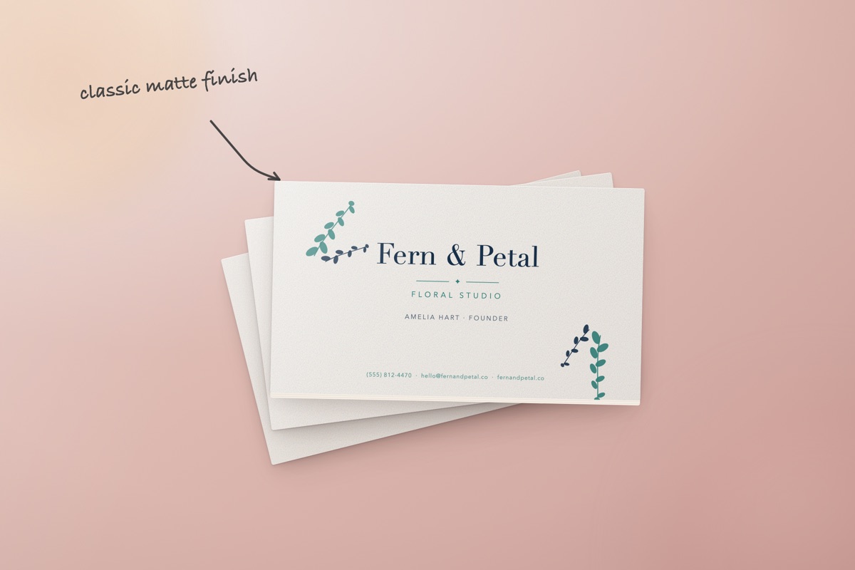 A fanned stack of matte business cards on a dusty pink surface, the top card printed with a botanical design for Fern and Petal Floral Studio, with a handwritten note and arrow reading "classic matte finish"